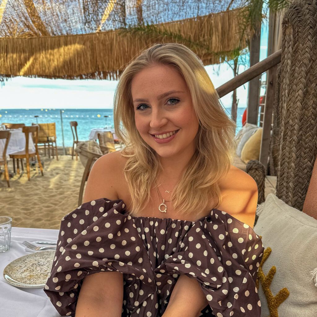 Vrouw met blonde haren draagt een Femme V. ketting met initialen op een strandlocatie in de zon